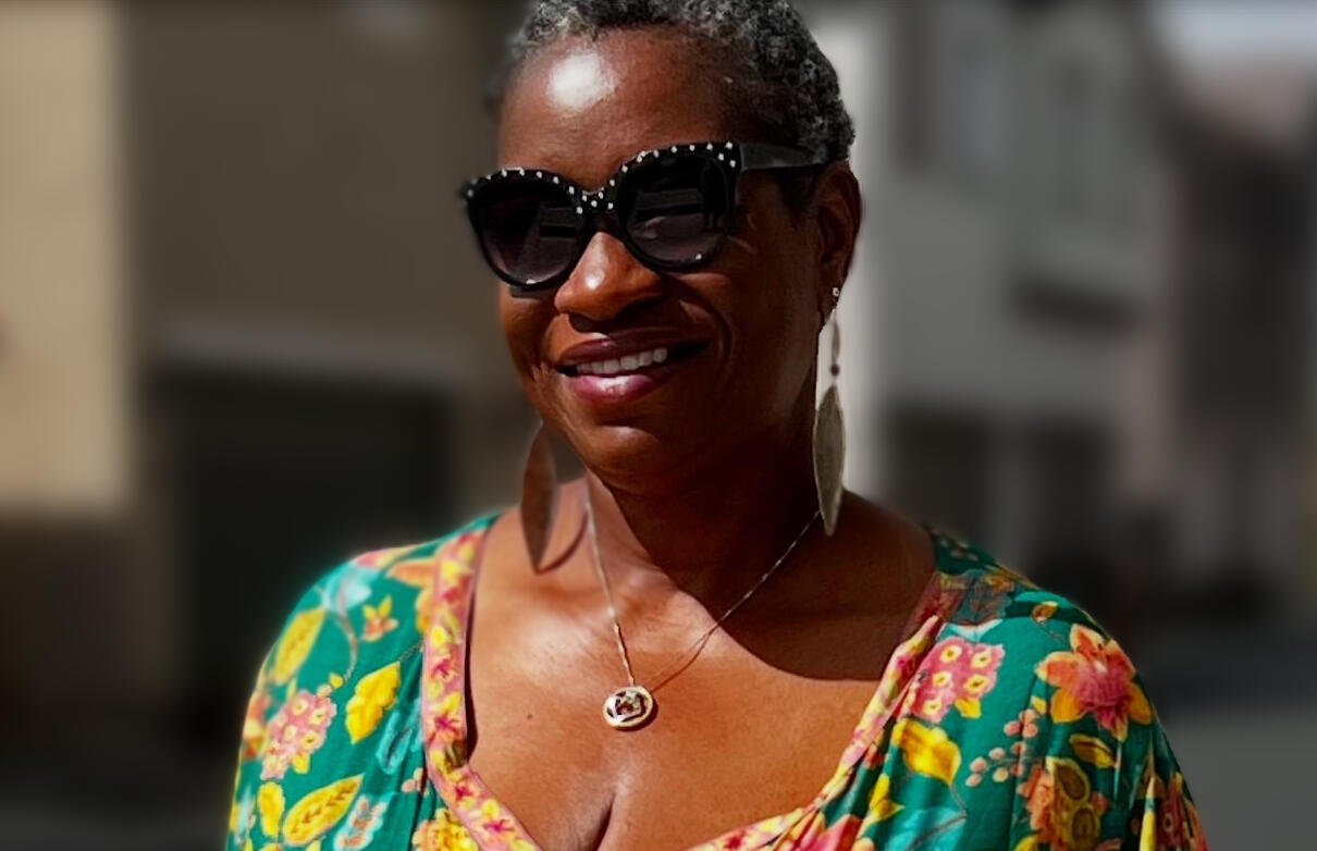 A smiling Black woman with short gray hair wearing large black sunglasses with decorative studs or rhinestones along the frames. She's dressed in a vibrant floral-patterned top or dress featuring bright colors including teal, yellow, pink, and green flower