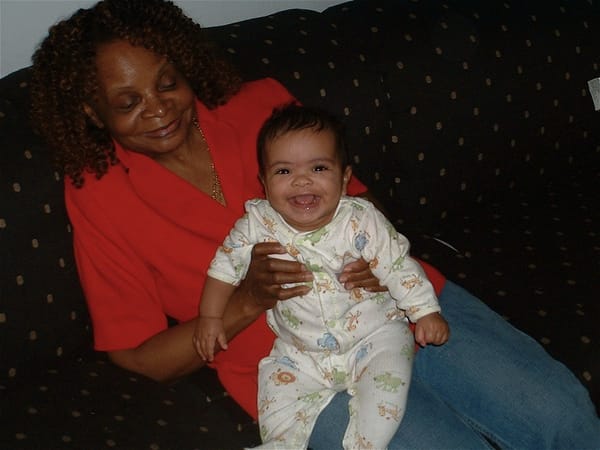 An older Black Woman, wearing a red shirt, blue jeans, and sitting atop a blue sofa, holds a baby on her lap. Both the baby a